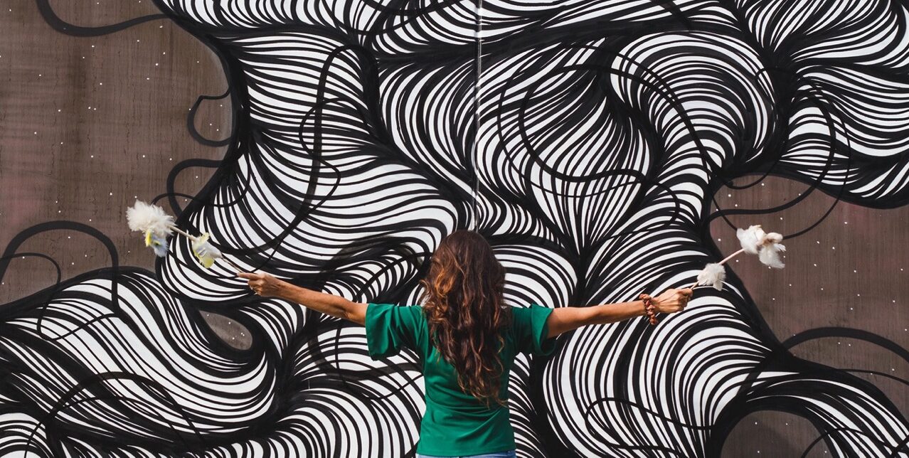 Serpent Mound Retreat - Evangeline Hemrick back turned with feather wands in each hand outstretched arms and zebra swirly background painted on brown concrete wall